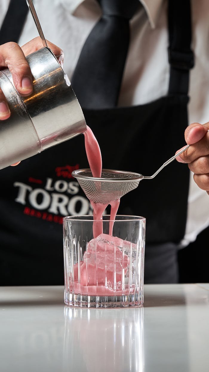 Bartender pours pink cocktail through strainer into glass with ice at upscale restaurant.